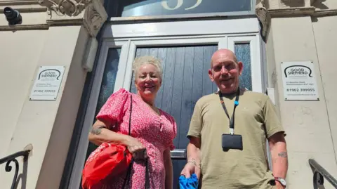 Good Shepherd A woman with blonde hair wearing a pink dress and a bald man in a beige t-shirt smile at the camera in front of the door of a building. There are signs next to the door saying 'Good Shepherd'.