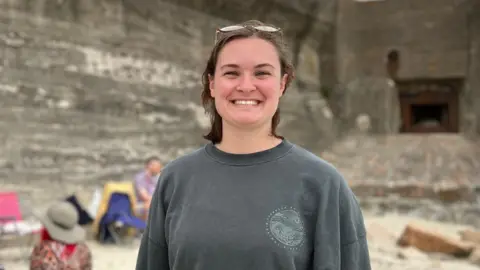 Kira is standing on a beach in front of a large concrete wall, wearing a dark long-sleeve top. A small group sits behind her on blankets and chairs, slightly out of focus.