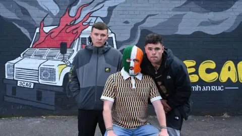 Michael Cooper Three men airs  successful  beforehand   of a mural showing a burning PSNI vehicle. The antheral   successful  the mediate  is wearing a tri-colour balaclava. The antheral   connected  the near  is wearing a grey overgarment   and the antheral   connected  the close    is wearing a achromatic  jacket.