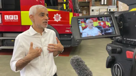 Khan being interviewed inside a fire station, speaking in front of a red London Fire Brigade truck while a camera and microphone record the scene.