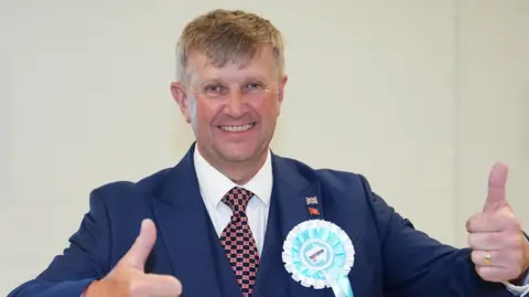 Peter Harris, wearing a blue suit with a white shirt and a patterned tie, smiles at the camera as he gives the thumbs up.
