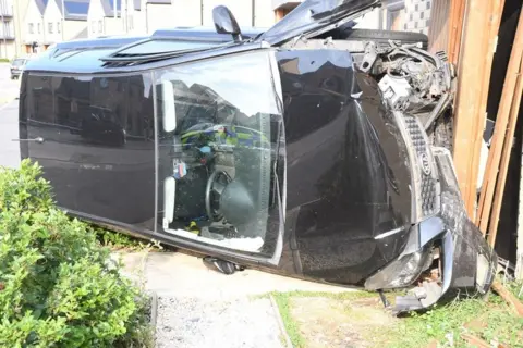 A black Kia car resting on the driver's side after it appears to have crashed into a house. The front of the car is all smashed up and the passenger door is slightly open.