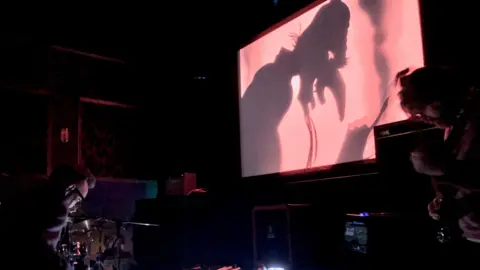 Flights of Helios A man hunched over a drum kit and another man playing a string instument play music in a dark room. A screen is above them showing an animated film, with the film showing a cartoon of a man with a long nose and beard looking down.