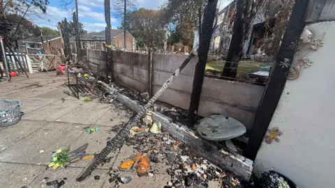 A fire-damaged garden is shown with charred fence posts, burnt debris and damaged outdoor furniture scattered across a patio area. In the background, several homes display extensive fire damage, including blackened walls and destroyed sections of the buildings. Among the wreckage are household ornaments, a bench and scorched garden features, illustrating the impact of the blaze on both the properties and surrounding outdoor spaces.