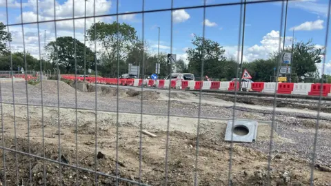 Julia Gregory / BBC A work site with metal fencing blocking it off. A mound of dirt can be seen on one side. Behind the works, a red and white plastic fence separates the site from a road, with cars on it.