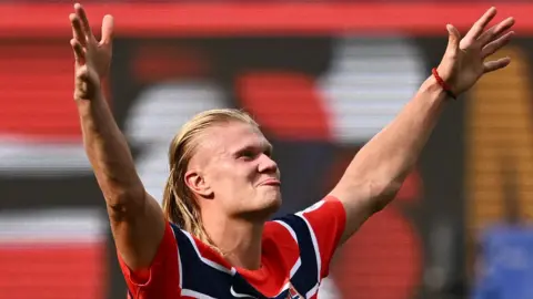 Erling Haaland with his arms in the air and looking happy and confident. He has long blonde hair and is wearing a red and blue striped Norway top. 