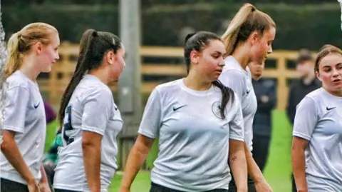 A close-up of five women on a football pitch. They are all wearing white football shirts. 