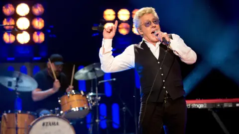 PA Media Roger Daltrey, a man with short greying hair and tinted glasses, is singing into a microphone while pointing upwards. He is wearing a black waistcoat and trousers with a white collared shirt. A man is playing the drums behind him. There are also stage lights and a keyboard in the background.
