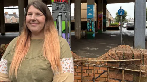 A woman with long blonde hair wearing a green top smiling in front of a wall and pillars that hold up the M4 with a road going under the motorway road deck