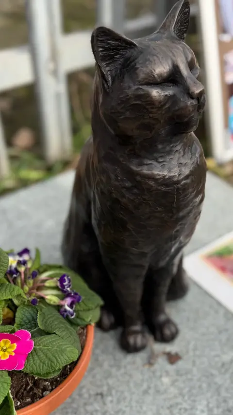 A black metal statue of a cat, sitting on a piece of granite beside a flowerpot.