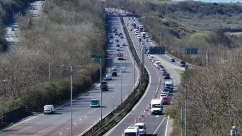 A two carriage A road. The carriageway on the left has one lane closed by cones and congested traffic. 