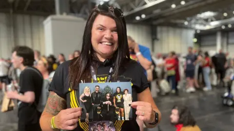 A woman is wearing a black t-shirt that says 'Back to the Beginning'. She is holding a photograph with the Osbourne family and smiling.