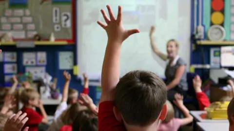 A rear view of schoolchildren raising their hands with a teacher in front of them raising her hand