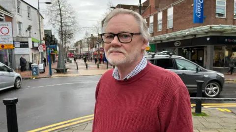 A man wearing a red jumper standing in front of a high street with cars going by.