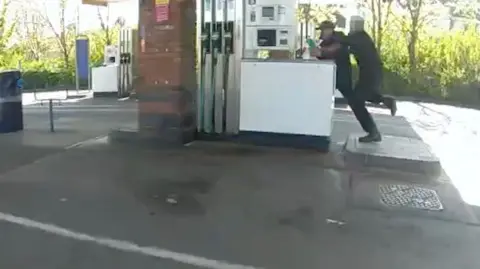 cctv footage of a petrol station forecourt. In the top right corner a police officer can be seen tackling a man doused in petrol holding a lighter