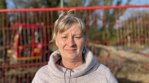 George Carden / BBC A woman in a pink jumper stood in front of a building site with a red fence