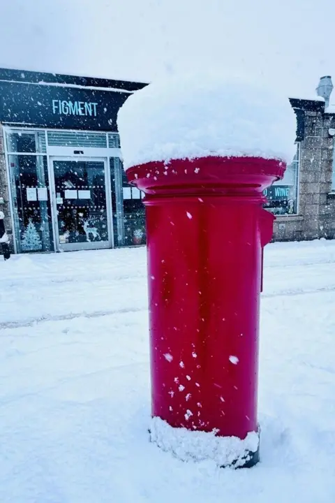 Lisa Megan A red mail box in deep snow with a lot of snow on its top, and a shop called Figment in the background.