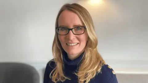 Sarah Wright A woman with shoulder-length blonde hair smiling at the camera. She is wearing a navy ribbed polo-neck jumper with gold buttons on the shoulder and black rimmed glasses. She is standing in a room with beige walls.