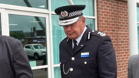 Kate Bradbrook/BBC A man dressed in a black police uniform walks with his head down past a brick building. 