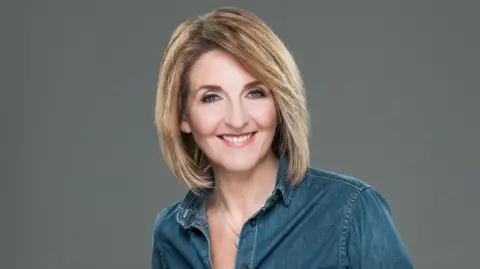 Kaye Adams smiling straight at the camera. She is wearing a blue shirt and is standing against a grey background.