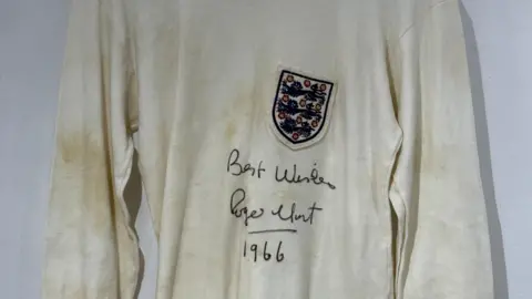 Pa/PA Wire A faded white shirt which has an England crest and 'best wishes, Roger Hunt, 1966' written in scrawled writing. The shirt also has brown mud stains on it.