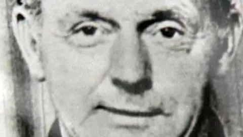 A cropped archive black and white picture showing Charles Armstrong. He is a middle aged man looking directly to camera wearing a dark jacket. Only his eyes and mouth are visible.