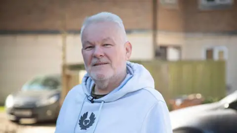 Un hombre que usa una sudadera gris con pelo gris y vello facial, de pie frente a una hilera de casas de ladrillo y autos estacionados