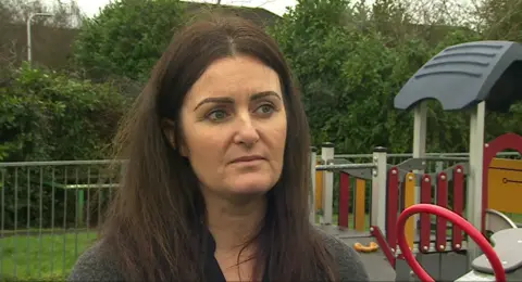 Joanna Zywica looking at the camera has brown hair, blue eyes and is wearing a grey cardigan. In the background is a children's playground.