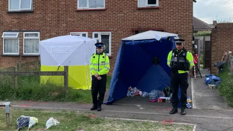 Sam Read/BBC Two police officers and a police tent outside a house. There are flowers laid on the ground and various items are under a blue tent