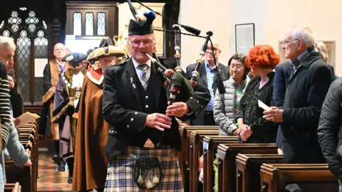 Reading Council A piper in traditional Scottish dress leading a procession of people down the aisle of a packed church