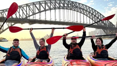 Family Handout Joe O'Sullivan is on the left of the picture wearing a light blue top and sitting in a kayak in front of the Sydney harbour Bridge in Australia. Leone Branski is on the right of the image, also in a kayak and wearing a black top. Both are holding their paddles aloft.
