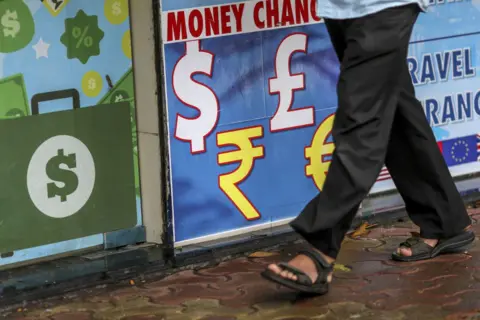Getty Images A wall with a money change board and a man walking past. Only his legs are seen.