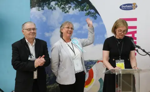 Chris Booth John Bailey, who has grey hair and glasses, wears a black suit and is clapping. His is standing next to Louise Taylor who is waving. She has short grey hair and wearing a light coloured top. She is smiling. They are next to a woman who is reading out election results.