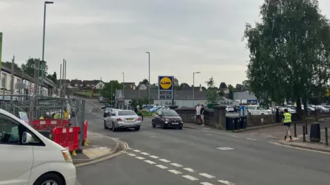 A silver Mercedes and a dark purple Fiat 500 drive past one another in a small town. There is construction to the left side of the bridge and a white car is trying to pull out of a junction near the construction works. A Lidl supermarket can be seen on the other side of the road. It is an overcast but hot day.