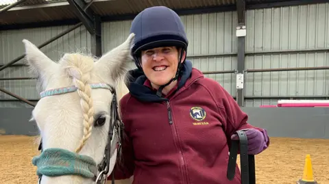 BBC Vanessa Tomas smiling at the camera and stood to the right of a white horse, which is white. Vanessa is wearing a burgandy jumper with the Erme Valley for the Disabled badge on it.