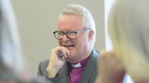 Diocese of Lincoln The Bishop of Grimsby, wearing a tweed jacket and traditional purple shirt, talks to two people who have their backs turned to the camera. He is smiling and has his hand on his chin.