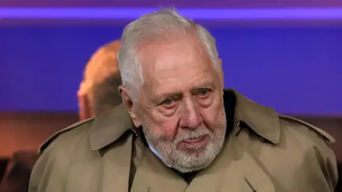 PA Media A man with short white hair and beard wearing a large brown mac coat