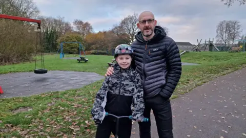 Matt, who is wearing a black puffer jacket and black jeans, is wearing glasses with his hand in his pocket, smiling into the camera with his hand on Charlie's shoulder. Charlie is wearing a helmet and camouflaged black and white coat and is on a scooter. There is an empty play area in the background with a few swings and a bench.