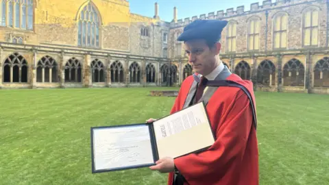 BBC Matt Baker wearing a red graduation cloak, on top of a grey blazer with a maroon tie, holding up a two sided folder which holds his certificate and documents. 
