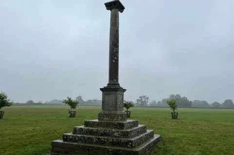 A monument dedicated to Percy Pilcher stands in a field with trees in the background.