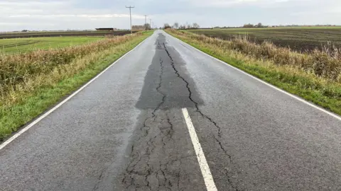 John Devine/BBC A long straight fen road, no street lights, minimal white lines, farmers fields either side of the road, that has large cracks running down the middle of it.