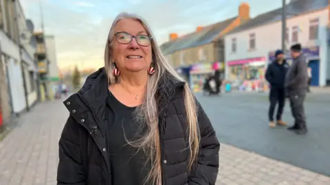 Jan Massey is smiling at the camera. She has long, straight, white hair and glasses. She is wearing a black puffer jacket over a black top. Her necklace has a pearl pendant and her red and white earrings resemble a Christmas tree bauble. She is standing in Front Street in Stanley. There are two men nearby. 