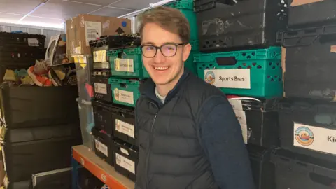 An indoor storage or warehouse‑like space filled with stacked plastic crates and boxes. A man wearing glasses is standing near a tall shelving unit filled with labelled crates. He is wearing a dark quilted gilet over a long‑sleeved navy top.