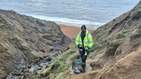 Badger Trust Isle of Wight Person in high-vis coat standing at top of cliff with green cage at their feet and badger inside.