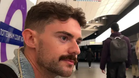 BBC Dan Harry, a young man with a moustache and wearing a denim jacket, is pictured sitting on a bench at Tottenham Court Road Tube station. He gazes off camera and is facing the tracks. Other people walk on by