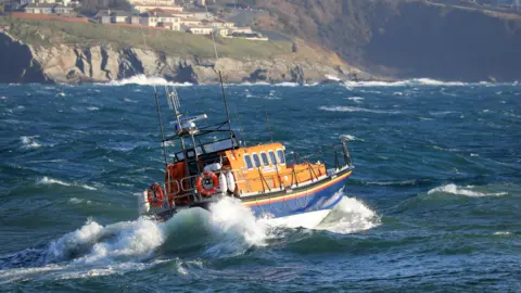 Douglas lifeboat sailing in a choppy sea. The boat is in the RNLI blue, orange and white colours and is leading to the left. Buildings can be seen on a headland in the background.