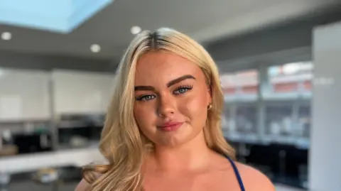 BBC The young women has long, blonde hair falling over her right shoulder. She is wearing a blue dress with thin straps and gold hoop earrings. She has tanned skin and blue eyes and is looking straight into the camera with a closed-mouth smile.