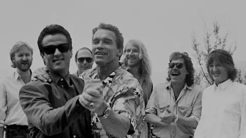 Richard Blanshard A black and white image of Arnold Schwarzenegger and Sylvester Stallone embracing one another as if about to dance together. Stallone is wearing sun glasses and a jacket and shirt. Schwarzenegger is wearing a printed shirt and watch. There are five people standing behind them laughing at the joke. 