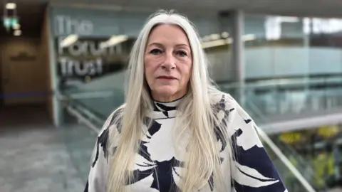 Paul Moseley/BBC Kay Mason Billig has long light-coloured hair and is wearing a patterned black-and-white top, standing indoors beside a glass balustrade. The softly blurred background shows a modern public building interior, with visible signage reading The Forum and bright natural lighting.