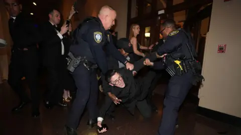 Niall Carson/PA Wire Police and security guards remove Isaac Burke from the National Building Museum. He is wearing black trousers and a black coat and is carrying a mobile phone.  His arms and legs are outstretched as he is lifted towards the door. Photographers are taking photos of Mr Burke in the background.